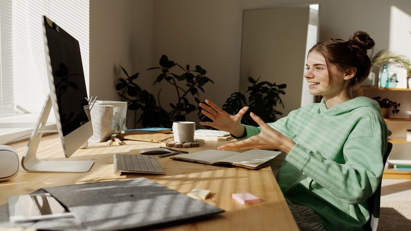 Person sitting at a desk in front of a computer, gesturing with both hands. The desk has a keyboard, mouse, notebook, coffee cup, and stationery. Plants and sunlight are in the background.